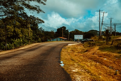 A rural road curves gently past a blue and orange house, surrounded by lush green trees on the left and an open grassy area on the right. A large sign welcomes visitors to Belize. Several power lines and a single vehicle are visible near the sign.