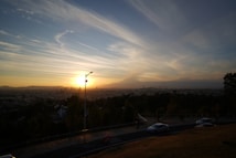 The scene features a dramatic sunset over a cityscape with a sprawling silhouette of buildings. The sky is filled with soft, wispy clouds that are beautifully lit by the warm glow of the setting sun. In the foreground, there is a road with a few cars and sparse figures, surrounded by trees.