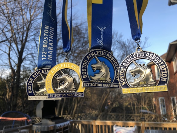 Three Boston Marathon medals are hanging against a backdrop of a clear blue sky and bare trees. Each medal features a unicorn emblem and the words 'Boston Athletic Association.' The medals are adorned with blue and yellow ribbons, showcasing different years of the marathon events.