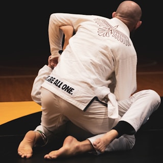 Two individuals are engaged in a jiu-jitsu match, wearing white gis. One person's back is prominently visible, showing an embroidered design and the phrase 'We Are All One.' The lighting is subdued, focusing on the competitors.
