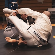 Two individuals are engaged in a Brazilian Jiu-Jitsu match on a mat. Both are wearing white gis and are in a close combat position, displaying intense focus and grappling skills. The setting appears to be an indoor sports facility with a wooden bench visible in the background.