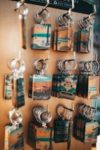 A display of multiple keychains on a wooden board. Each keychain features a rectangular plate with vintage-style illustrations related to California, including images of beaches, bicycles, and iconic routes. The keychains have a rustic and nostalgic look, highlighting travel and tourism themes.