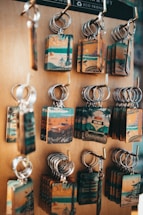 A display of multiple keychains on a wooden board. Each keychain features a rectangular plate with vintage-style illustrations related to California, including images of beaches, bicycles, and iconic routes. The keychains have a rustic and nostalgic look, highlighting travel and tourism themes.