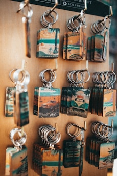 A display of multiple keychains on a wooden board. Each keychain features a rectangular plate with vintage-style illustrations related to California, including images of beaches, bicycles, and iconic routes. The keychains have a rustic and nostalgic look, highlighting travel and tourism themes.