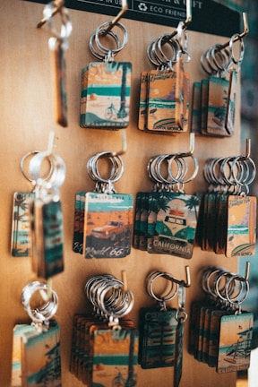 A display of multiple keychains on a wooden board. Each keychain features a rectangular plate with vintage-style illustrations related to California, including images of beaches, bicycles, and iconic routes. The keychains have a rustic and nostalgic look, highlighting travel and tourism themes.