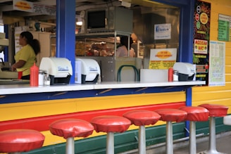 A brightly colored food counter with a retro design features a row of red-cushioned stools in front. Behind the counter, various kitchen equipment and supplies are visible, including a microwave and stacked bread. Two people appear to be working behind the counter. The counter displays condiment dispensers, napkin holders, and promotional signs on the wall advertising menu items and specials.