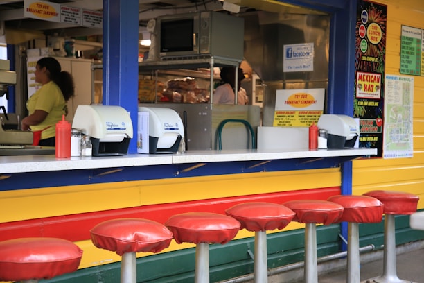 A brightly colored food counter with a retro design features a row of red-cushioned stools in front. Behind the counter, various kitchen equipment and supplies are visible, including a microwave and stacked bread. Two people appear to be working behind the counter. The counter displays condiment dispensers, napkin holders, and promotional signs on the wall advertising menu items and specials.