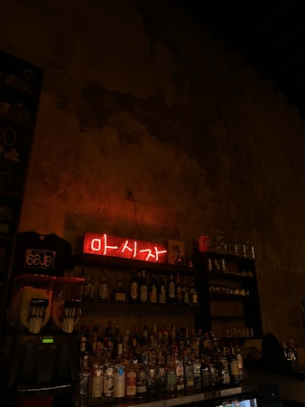 A dimly lit bar setting with shelves stocked with various liquor bottles. A bright red neon sign with characters in another language hangs above the bottles. The atmosphere is rustic, with a textured, unfinished wall in the background. To the left, there is a frozen drink dispenser and a t-shirt with a logo displayed.
