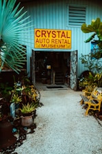 An entrance to a museum labeled 'Crystal Auto Rental Museum' with a large yellow and red sign. The doorway is framed with decorative elements and surrounded by lush plants and potted greenery. A path made of small white stones leads to the entrance, flanked by yellow painted benches. The building exterior is a muted blue with ventilation panels above the entrance.