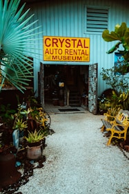 An entrance to a museum labeled 'Crystal Auto Rental Museum' with a large yellow and red sign. The doorway is framed with decorative elements and surrounded by lush plants and potted greenery. A path made of small white stones leads to the entrance, flanked by yellow painted benches. The building exterior is a muted blue with ventilation panels above the entrance.