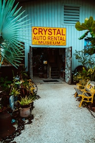 An entrance to a museum labeled 'Crystal Auto Rental Museum' with a large yellow and red sign. The doorway is framed with decorative elements and surrounded by lush plants and potted greenery. A path made of small white stones leads to the entrance, flanked by yellow painted benches. The building exterior is a muted blue with ventilation panels above the entrance.
