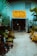 An entrance to a museum labeled 'Crystal Auto Rental Museum' with a large yellow and red sign. The doorway is framed with decorative elements and surrounded by lush plants and potted greenery. A path made of small white stones leads to the entrance, flanked by yellow painted benches. The building exterior is a muted blue with ventilation panels above the entrance.