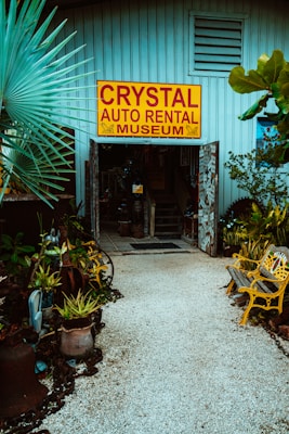 An entrance to a museum labeled 'Crystal Auto Rental Museum' with a large yellow and red sign. The doorway is framed with decorative elements and surrounded by lush plants and potted greenery. A path made of small white stones leads to the entrance, flanked by yellow painted benches. The building exterior is a muted blue with ventilation panels above the entrance.