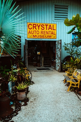 An entrance to a museum labeled 'Crystal Auto Rental Museum' with a large yellow and red sign. The doorway is framed with decorative elements and surrounded by lush plants and potted greenery. A path made of small white stones leads to the entrance, flanked by yellow painted benches. The building exterior is a muted blue with ventilation panels above the entrance.