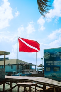 A bright red flag with a white diagonal stripe is prominently displayed against a backdrop of a vibrant blue sky with scattered clouds. Below, there is a wooden dock leading to a building labeled 'AMIGOS DEL MAR'. To the right, signage for 'Ocean Ferry' details boat departure times and destinations. Palm leaves are partially visible at the top right, suggesting a coastal or tropical setting.