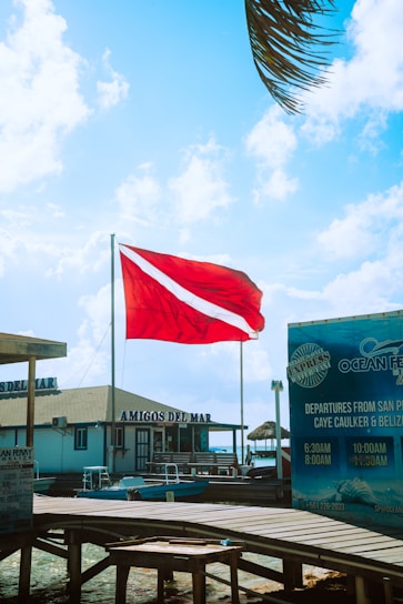 A bright red flag with a white diagonal stripe is prominently displayed against a backdrop of a vibrant blue sky with scattered clouds. Below, there is a wooden dock leading to a building labeled 'AMIGOS DEL MAR'. To the right, signage for 'Ocean Ferry' details boat departure times and destinations. Palm leaves are partially visible at the top right, suggesting a coastal or tropical setting.