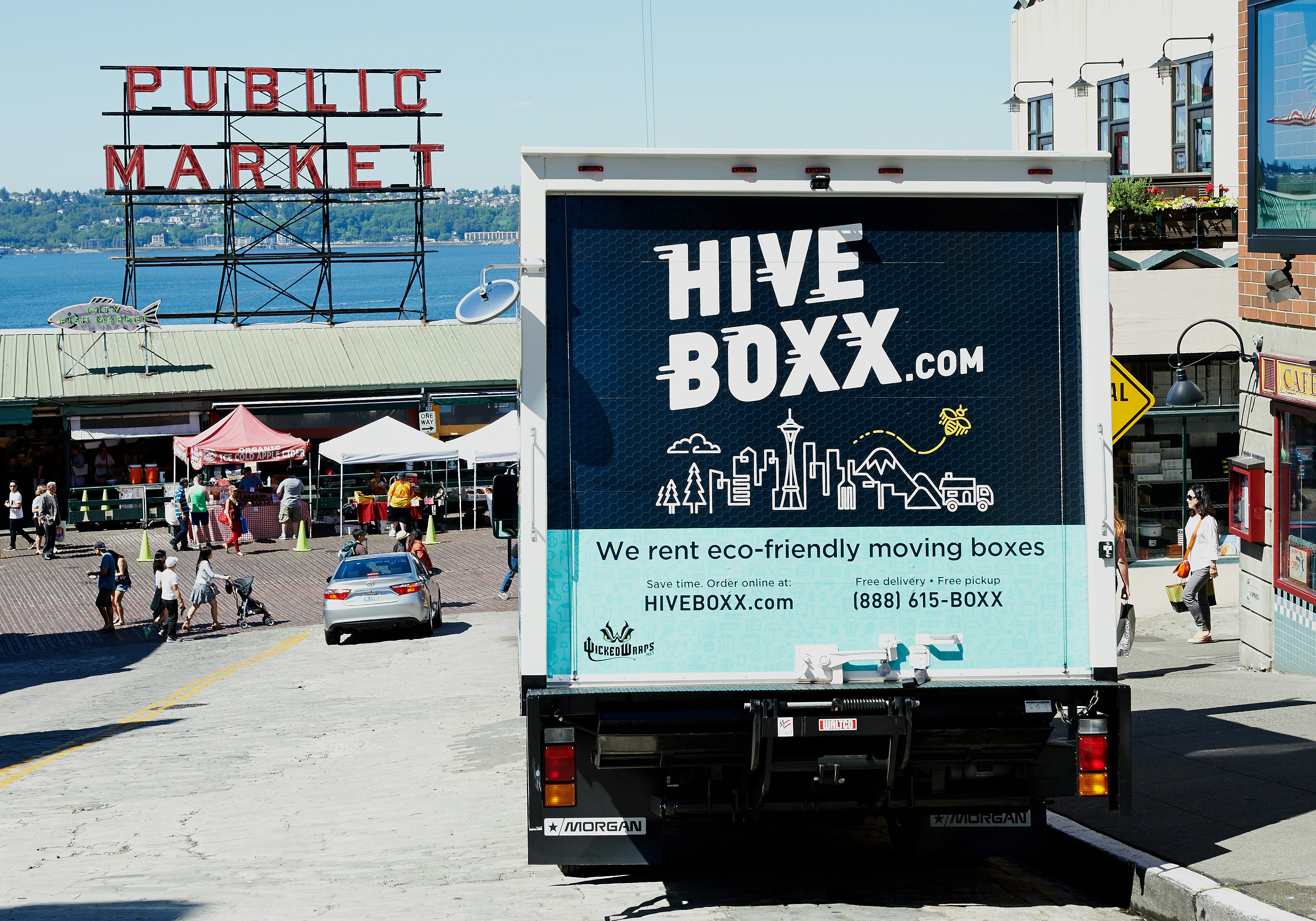 A truck with a 'HIVE BOXX' advertisement is parked on a sloped street near a public market, with a view of the water and distant hills. People are walking around, and several market stalls with tents are visible. The truck promotes eco-friendly moving boxes with a scenic city graphic on the back, featuring iconic landmarks and natural elements.