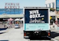 A truck with a 'HIVE BOXX' advertisement is parked on a sloped street near a public market, with a view of the water and distant hills. People are walking around, and several market stalls with tents are visible. The truck promotes eco-friendly moving boxes with a scenic city graphic on the back, featuring iconic landmarks and natural elements.