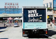 A truck with a 'HIVE BOXX' advertisement is parked on a sloped street near a public market, with a view of the water and distant hills. People are walking around, and several market stalls with tents are visible. The truck promotes eco-friendly moving boxes with a scenic city graphic on the back, featuring iconic landmarks and natural elements.