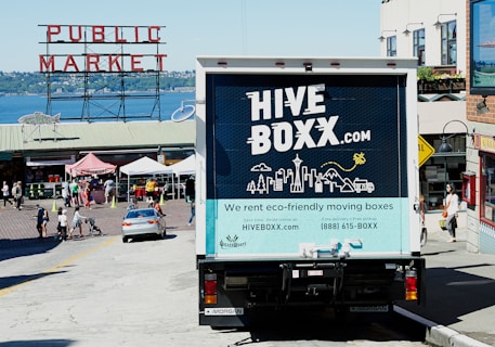 A truck with a 'HIVE BOXX' advertisement is parked on a sloped street near a public market, with a view of the water and distant hills. People are walking around, and several market stalls with tents are visible. The truck promotes eco-friendly moving boxes with a scenic city graphic on the back, featuring iconic landmarks and natural elements.