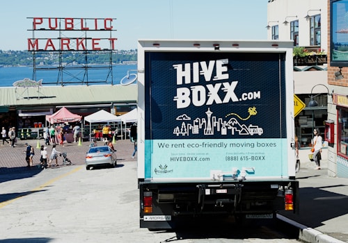 A truck with a 'HIVE BOXX' advertisement is parked on a sloped street near a public market, with a view of the water and distant hills. People are walking around, and several market stalls with tents are visible. The truck promotes eco-friendly moving boxes with a scenic city graphic on the back, featuring iconic landmarks and natural elements.