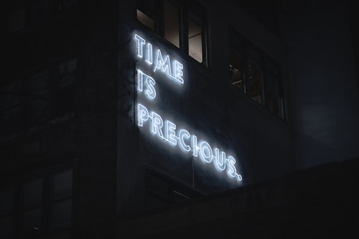 A neon sign on a building displays the words 'TIME IS PRECIOUS.' in bright white letters. The scene is dark, and the sign stands out against the dimly lit windows and the shadowy facade of the building. The atmosphere feels quiet and contemplative.