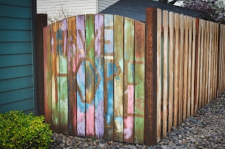 A wooden fence with an arched gate displays colorful graffiti with the words 'Have Hope'. The diverse hues of paint contrast with the natural wood texture. The setting includes a small green shrub on the left and gravel ground extending to the right.