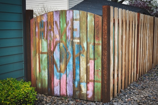 A wooden fence with an arched gate displays colorful graffiti with the words 'Have Hope'. The diverse hues of paint contrast with the natural wood texture. The setting includes a small green shrub on the left and gravel ground extending to the right.