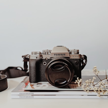 A vintage-style camera with a black body and silver top is resting on a stack of magazines titled 'Vision Magazine'. A camera strap is partially visible on the left side, and a small sprig of delicate white flowers lies to the right of the camera, adding a touch of elegance.