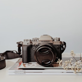 A vintage-style camera with a black body and silver top is resting on a stack of magazines titled 'Vision Magazine'. A camera strap is partially visible on the left side, and a small sprig of delicate white flowers lies to the right of the camera, adding a touch of elegance.