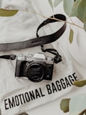 A Fujifilm camera with a black strap rests on a white fabric that has the text 'Emotional Baggage' printed on it. Green leaves from a plant are partially visible, adding a natural element to the composition.