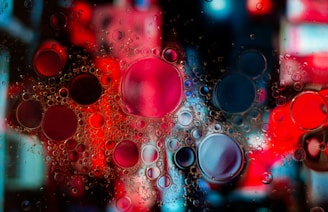 A vibrant and abstract composition of oil droplets suspended in water with a colorful background. The droplets vary in size and create a pattern of overlapping circles. The interplay of light gives the image a dynamic and lively appearance.