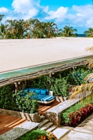 A serene outdoor lounge area with a curved blue sectional sofa set amidst lush green plants. The space is accented by stone walls and wooden decking with stairs leading down. Palm trees are visible in the background against a bright blue sky.