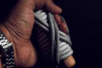 A hand gripping a coiled rope with a wooden handle, wearing a silver metallic wristwatch. The rope is wrapped tightly around the handle, creating a textured appearance. The lighting is dim, casting a shadowy effect on the scene.