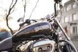 A close-up view of a Harley-Davidson motorcycle with a focus on the fuel tank and engine, set against an urban background with blurred buildings and light peeking through tree branches.