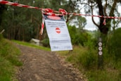 A dirt path in a forested area is blocked by red and white caution tape with a sign that states the closure of the 1000 Steps/Kokoda track and Lyrebird Track until further notice. The sign is attached to the tape, and surrounding the path are green trees and bushes.