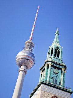 A tall, modern television tower featuring a silver sphere with antennas stands beside a historic tower with a green, ornate spire and a clock. The clear blue sky creates a vivid backdrop.