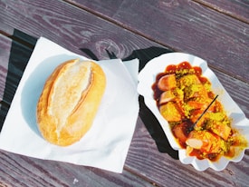A bread roll is placed on a white napkin next to a tray filled with sliced sausage covered in curry sauce and sprinkled with yellow curry powder. The tray is set on a wooden surface, and there's a toothpick or small fork in the tray.