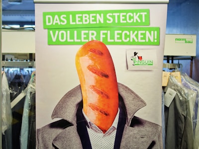 A poster features a humorous image of a person with a loaf of bread as a head. The person is dressed in a suit, and the background is busy with racks of clothing covered in plastic. The text on the poster is in German and translates to 'Life is full of stains!'. There is a small logo with a penguin included.