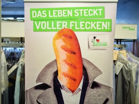 A poster features a humorous image of a person with a loaf of bread as a head. The person is dressed in a suit, and the background is busy with racks of clothing covered in plastic. The text on the poster is in German and translates to 'Life is full of stains!'. There is a small logo with a penguin included.