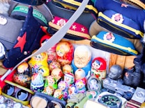 A collection of colorful Russian nesting dolls, various Soviet-era military hats, and vintage items including binoculars and cameras arranged closely together. The combination of traditional and military-themed objects creates a sense of nostalgia and history.
