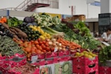 A vibrant market stall displays a variety of fresh vegetables, including tomatoes, carrots, onions, cucumbers, peppers, zucchinis, green beans, and leafy greens. The produce is arranged in piles over pink cardboard boxes with labels. The setting is an indoor market with a person in the background.