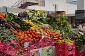 A vibrant market stall displays a variety of fresh vegetables, including tomatoes, carrots, onions, cucumbers, peppers, zucchinis, green beans, and leafy greens. The produce is arranged in piles over pink cardboard boxes with labels. The setting is an indoor market with a person in the background.