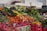 A vibrant market stall displays a variety of fresh vegetables, including tomatoes, carrots, onions, cucumbers, peppers, zucchinis, green beans, and leafy greens. The produce is arranged in piles over pink cardboard boxes with labels. The setting is an indoor market with a person in the background.