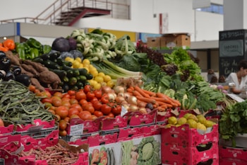A vibrant market stall displays a variety of fresh vegetables, including tomatoes, carrots, onions, cucumbers, peppers, zucchinis, green beans, and leafy greens. The produce is arranged in piles over pink cardboard boxes with labels. The setting is an indoor market with a person in the background.