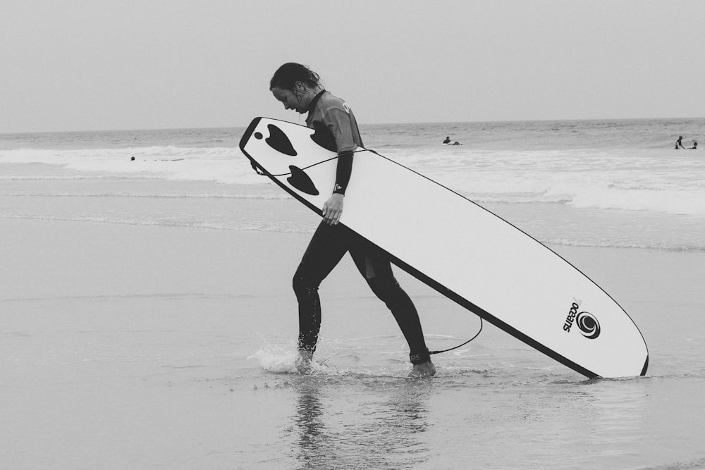 검은 잠수복을 입은 남자가 낮에 해변을 걷고 있는 흰색 서핑보드를 들고