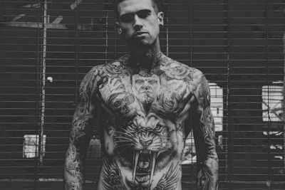 A person with extensive tattoos covering the chest and arms stands in front of a barred window. The tattoos include a large roaring tiger on the chest, skulls, human faces, and intricate designs covering the arms. The setting appears to be industrial with a moody atmosphere.