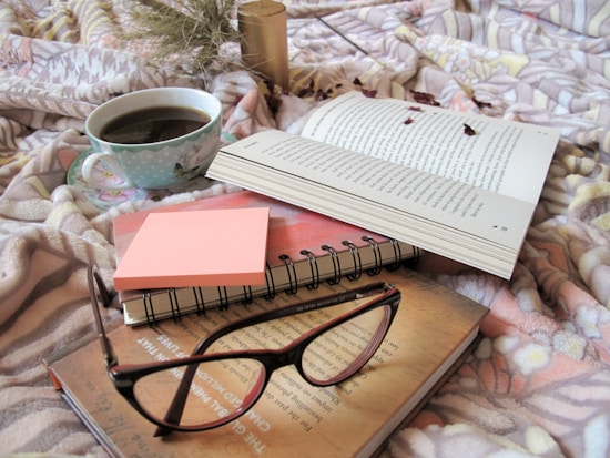 An open book rests on a pastel patterned blanket, surrounded by a warm cup of coffee and a stack of sticky notes. Nearby, a pair of eyeglasses lies atop a closed notebook.