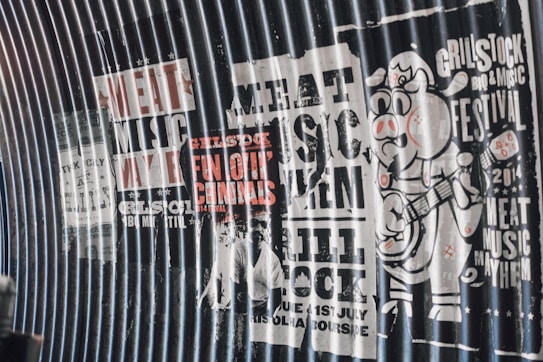 A series of vintage-style posters on a corrugated metal surface promoting a music and BBQ festival. The posters feature bold text with phrases like 'Meat Music Mayhem' and 'Grillstock BBQ & Music Festival 2014'. There is a cartoon pig character playing a guitar on the rightmost poster.