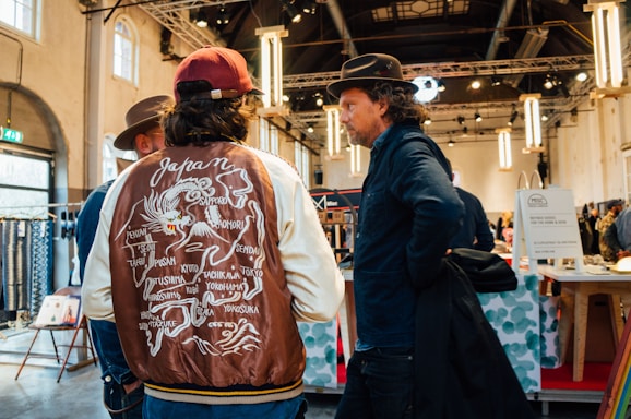 Three men are standing in a well-lit indoor space, possibly a market or showroom. One man is wearing a jacket with a map of Japan printed on the back, while the other two wear hats and casual attire. The setting includes large hanging lights and various booths and displays.